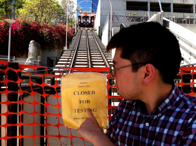 Los Angeles Downtown News reporter Eddie Kim points to the problem, as the two Angels Flight funicular railway cars hang in limbo at the top of the tracks to Bunker Hill.