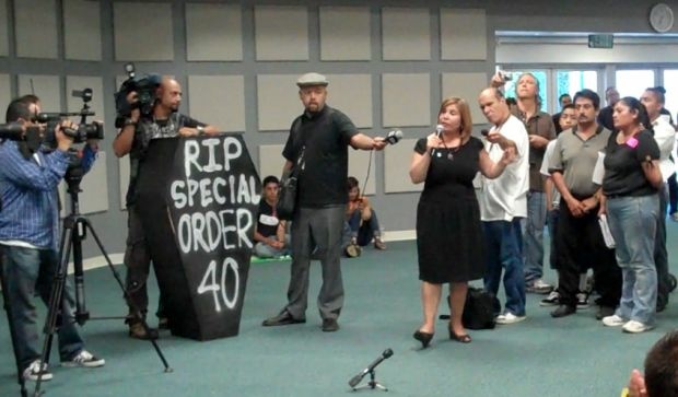 A camera operator rests on the mock coffin as Angelica Salas, executive director of the Coalition for Humane Immigrant Rights of Los Angeles, addresses a Homeland Security panel Monday, August 15, 2011