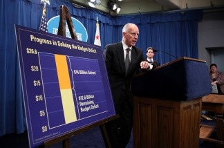 California Gov. Jerry Brown stands next to a chart that shows dollar amounts in the millions that were cut from the State's budget.