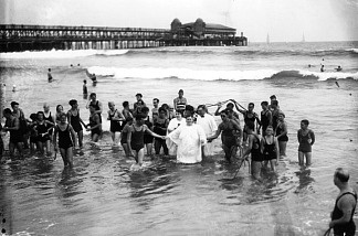 Mercer 5382 A woman wearing a white baptismal gown is led from the ocean by witnesses in bathing suits after her baptism in the surf next to the Municipal Pier in Long Beach.