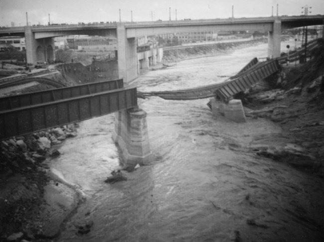 1938: People walk along the flooded L.A. River near the Dayton Avenue Bridge. In the background, a railroad bridge hangs in a twisted heap after one of the pilings has collapsed. (Photograph courtesy of the Los Angeles Public Library)