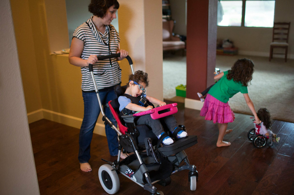 Kathleen helps Gideon into his chair while his sister Genevieve plays with her dolls, one of whom has a wheelchair.