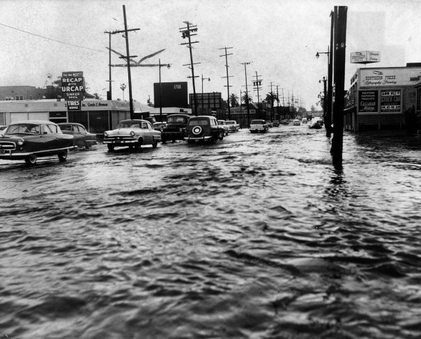 1938: People walk along the flooded L.A. River near the Dayton Avenue Bridge. In the background, a railroad bridge hangs in a twisted heap after one of the pilings has collapsed. (Photograph courtesy of the Los Angeles Public Library)