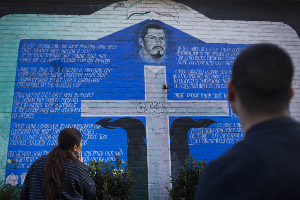 Ramona Gardens Police Relations - 2 Amanda Gutierrez, left, and Eddie Licon look at a mural in Ramona Gardens in Boyle Heights memorializing a community member killed by police in the 1990s.
