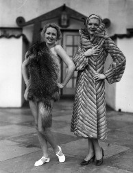 Mercer 5848 C. 1930: A fashion show including bathing suits, clothes for children, furs, and wedding dresses was held in a large Spanish-style courtyard. View 12: Young women standing next to each other model two distinctly different furs. The model on the right wears a long gray striped coat, tan hat, and black heels; the woman on the left appears to be bare underneath an animal pelt, possibly a fox fur pelt, which is draped over her right shoulder and around her hips, and wearing white shoes.