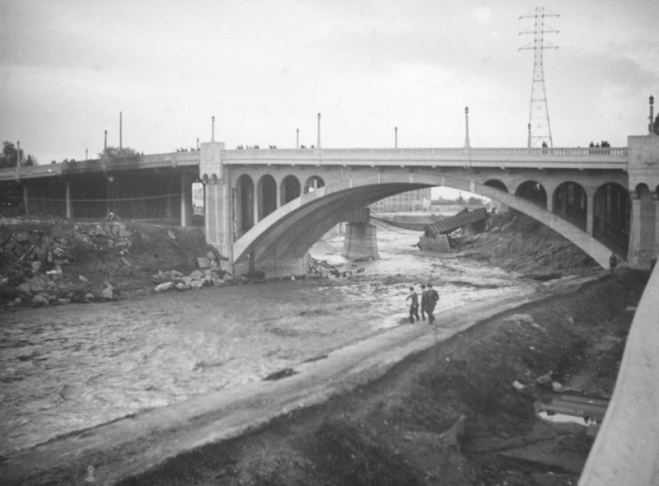 1938: People walk along the flooded L.A. River near the Dayton Avenue Bridge. In the background, a railroad bridge hangs in a twisted heap after one of the pilings has collapsed. (Photograph courtesy of the Los Angeles Public Library)