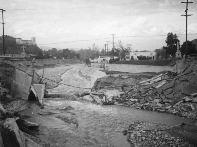 1938: People walk along the flooded L.A. River near the Dayton Avenue Bridge. In the background, a railroad bridge hangs in a twisted heap after one of the pilings has collapsed. (Photograph courtesy of the Los Angeles Public Library)
